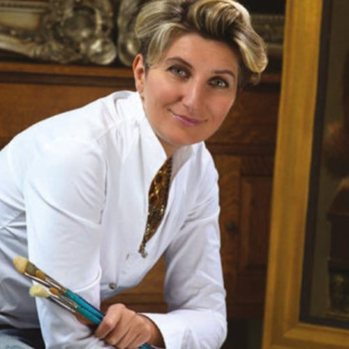 rofessional with blonde hair in white coat holding paintbrushes in art workshop with wooden features.